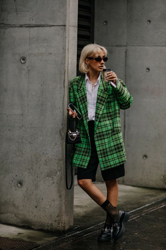 Foto para ilustrar 7 looks para usar essa semana. Uma mulher loira com cabelos curtos descoloridos está em frente a uma parede de concreto. O look é composto por camisa branca e bermuda de alfaiataria preta na altura do joelho, blazer oversized verde xadrez, míni bolsa preta, mocassim com solado tratorado preto envernizado e meia fio 15 na altura da canela.