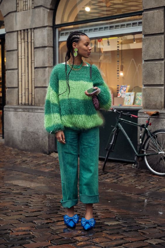 Foto para ilustrar ideias de looks para usar essa semana. Look de domingo. Uma mulher com cabelos trançados está em frente a uma livraria. O look é composto por um suéter peludinho listrado em tons de verde escuro e verde claro, calça larga verde escuro com a barra dobrada, brincos grandes verde e scarpin azul com laço.