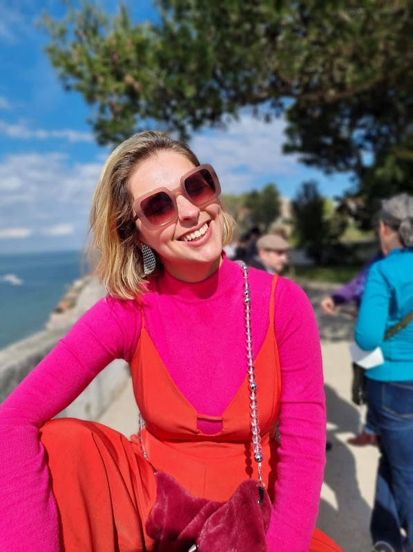 Foto destaque de Marcie sorrindo em uma paisagem natural ao fundo. O look é composto por óculos de sol quadrados rosa, blusa de gola alta fúcsia e macacão laranja.