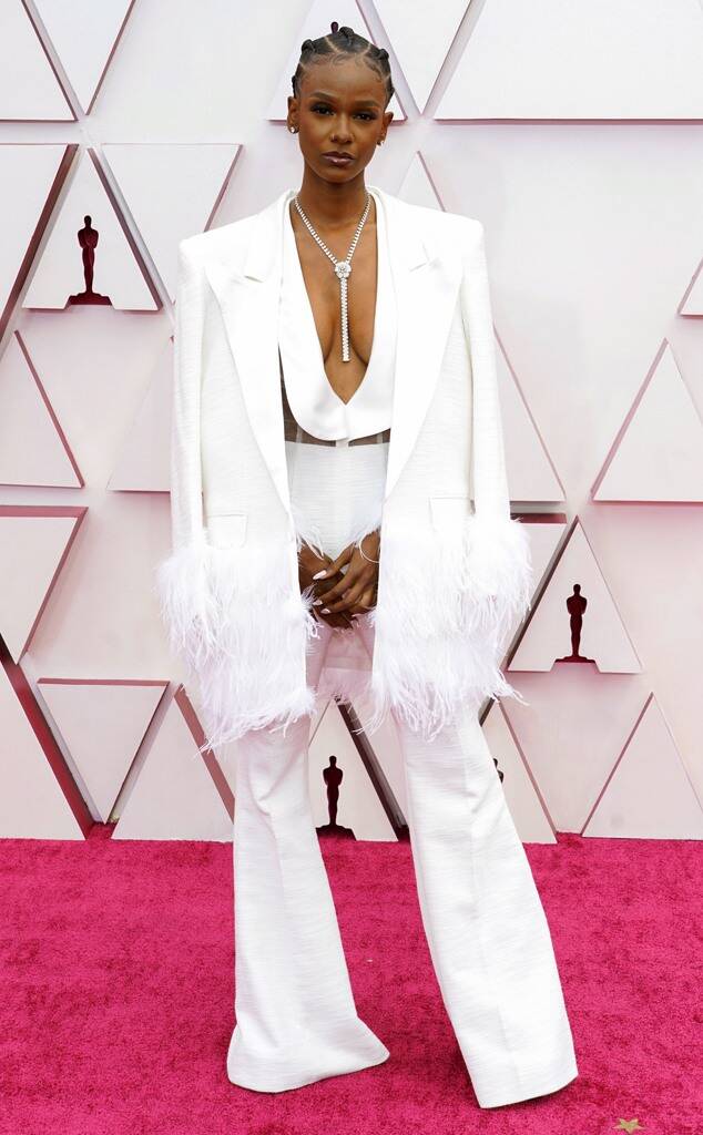 Foto com Tiara Thomas no red carpet do Oscar 2021. O look é composto por macacão branco decotado até o umbigo com lapelas largas e calça de alfaiataria, colar gravata prateado e blazer branco nos ombros, com mangas e barra com plumas. Os cabelos estão trançados para trás.