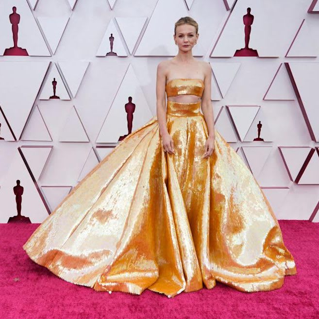 Foto mostrando o vestido de Carey Mulligan no Oscar 2021. Obra de Valentino, o look é composto por cropped sem alças de paetês dourados, saia de cintura alta longa volumosa no estilo princesa também de paetês dourados. Os cabelos estão presos em um coque baixo e a make é leve, com batom escuro.