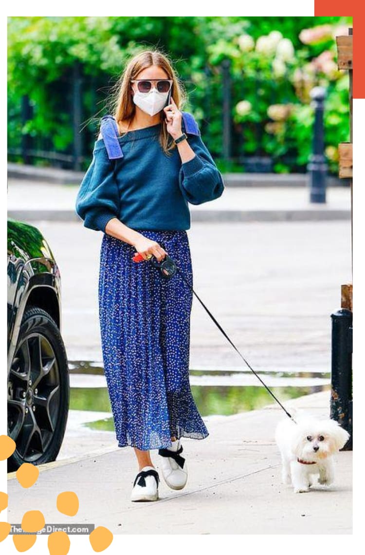 Foto vertical com borda branca e detalhe laranja. no topo direito. Na imagem, Olivia Palermo caminha na rua com seu cachorro em uma guia. O look é composto por moletom de mangas fluídas azul com decote ombro a ombro e alças largas em um tom de azul próximo ao índigo, saia mídi plissada azul com poás brancos, tênis branco com laço preto.
