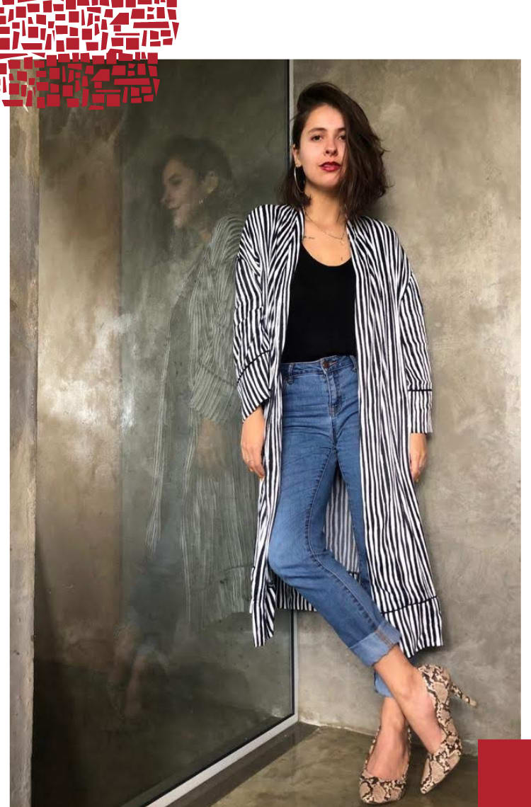 Foto vertical com moldura branca. Na foto, Marcie posa de pé com uma perna cruzada em frente a uma parede cinza. O look é composto por camiseta preta, quimono listrado preto e branco, jeans com barra dobrada e scarpin com estampa de cobra.