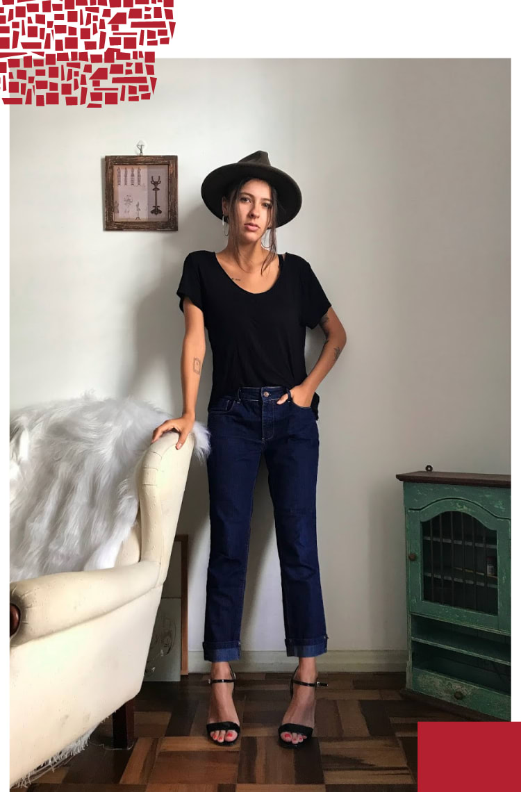 Foto vertical com moldura branca. Na foto, Marcie posa levemente escorada em uma poltrona branca com pelego branco. O look é composto por chapéu com cabelos presos e duas mechas só para a frente, camiseta preta com decote v, calça jeans boyfriend e sandália preta com tiras finas.