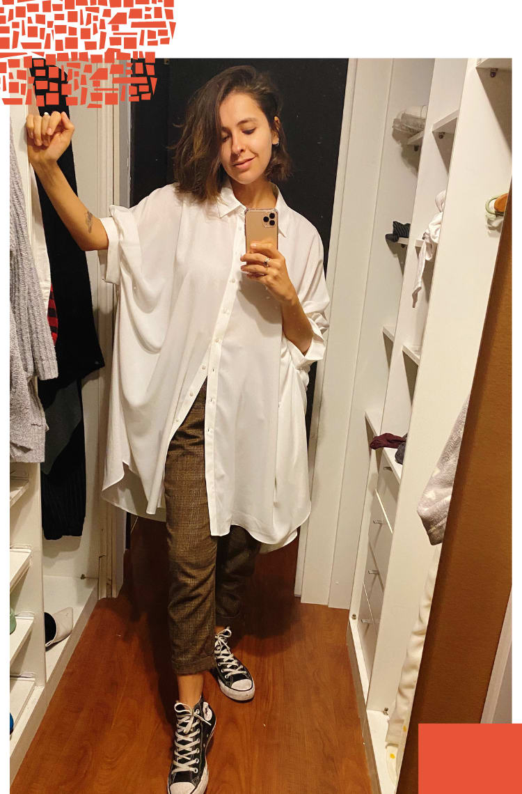 Foto vertical com moldura branca. Na imagem, Marcie (cabelos curtos e castanhos na altura do ombro) posa em frente a um espelho de closet, com o celular na mão. O look é composto por camisa oversized branca de mangas curtas, calça de alfaiataria xadrez marrom e all star botinha preto.