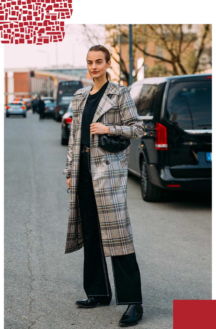 Imagem vertical com borda branca, na qual uma mulher com cabelos castanhos presos em um rabo caminha em uma rua com carros estacionados. O look é composto por camiseta preta com gola v, calça jeans preta, trench coat xadrez cinza.