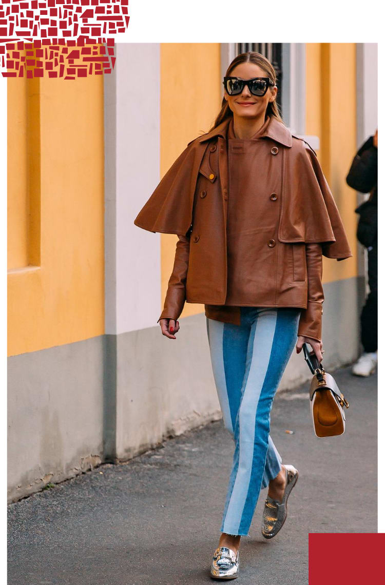 Foto vertical com moldura branca em volta. Na foto, Olivia Palermo caminha em uma rua vazia, com uma parede amarela ao fundo. O look é composto por uma jaqueta de couro marrom oversized com babados no ombro, calça jeans bicolor azul escuro com azul claro, bolsa marrom e loafer prateado.