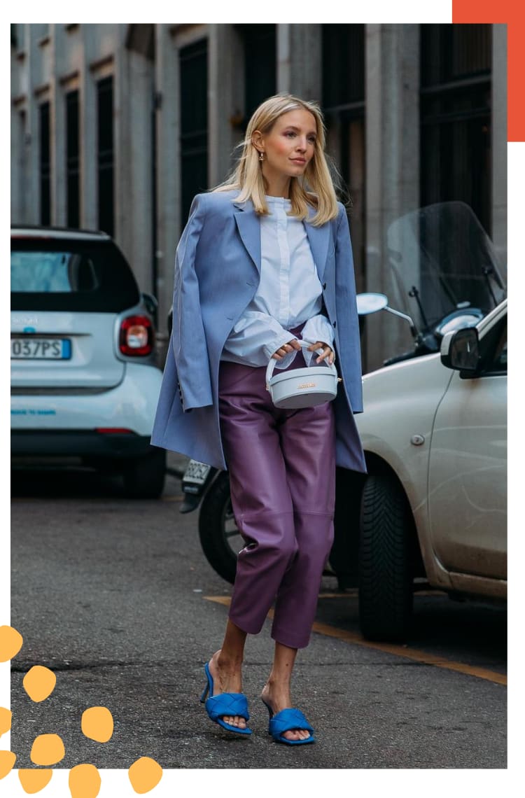 Imagem com moldura branca, na qual uma mulher loira com cabelos lisos. O look é composto por camisa branca, blazer lilás, calça violeta de couro, tamanco de couro roxo tressê.