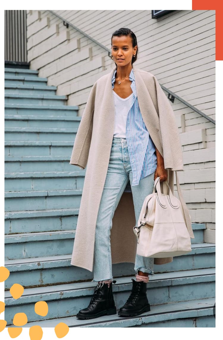 Foto vertical com moldura branca. Na foto, uma mulher para em uma escada azul e parede branca. O look é composto por camiseta branca com gola v, calça jeans, camisa listrada branca e azul, casaco de lã batida alongada bege, coturno preto, com meia laranja.