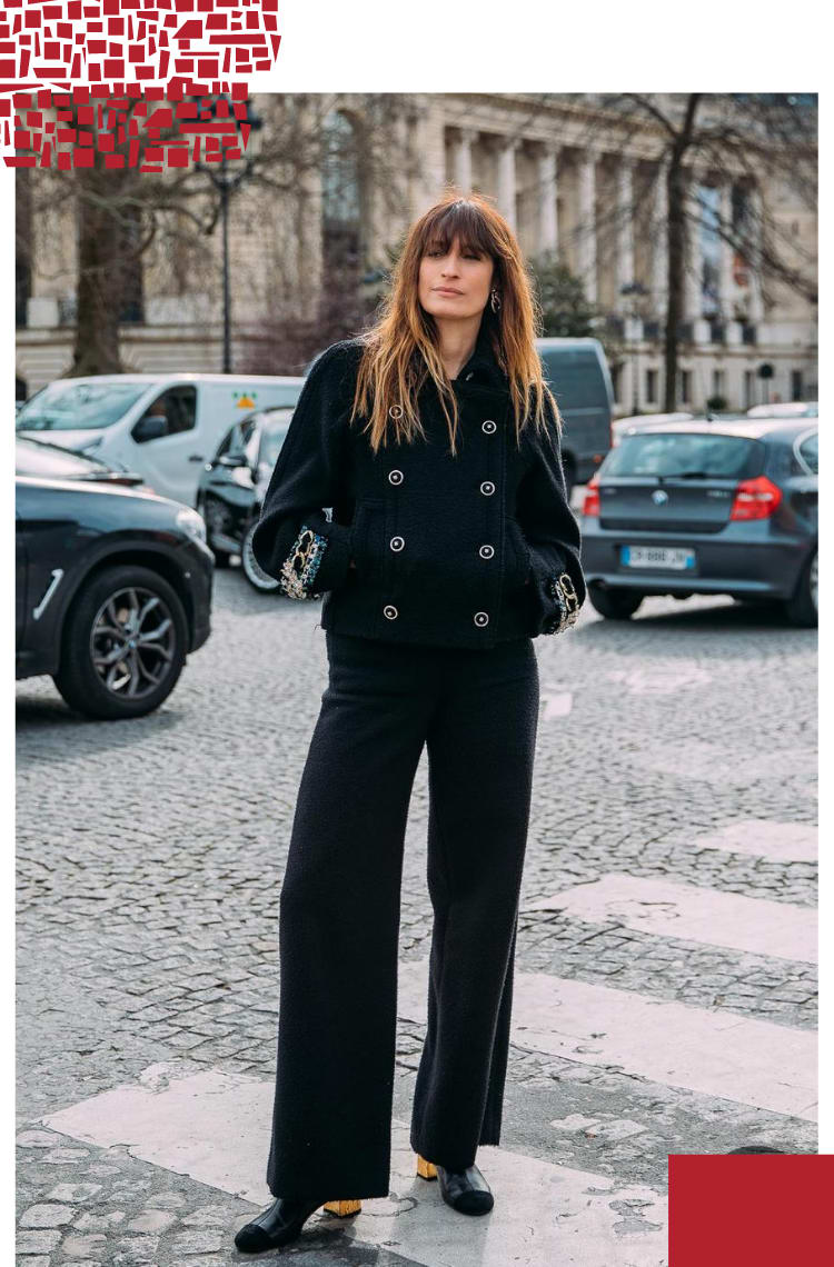 Foto vertical com moldura branca. Na foto, uma mulher com cabelos castanhos e franja está parada em frente de um estacionamento. O look é composto por jaqueta preta de tweed e botões dourados, calça de alfaiataria preta e bota preta com salto dourado.