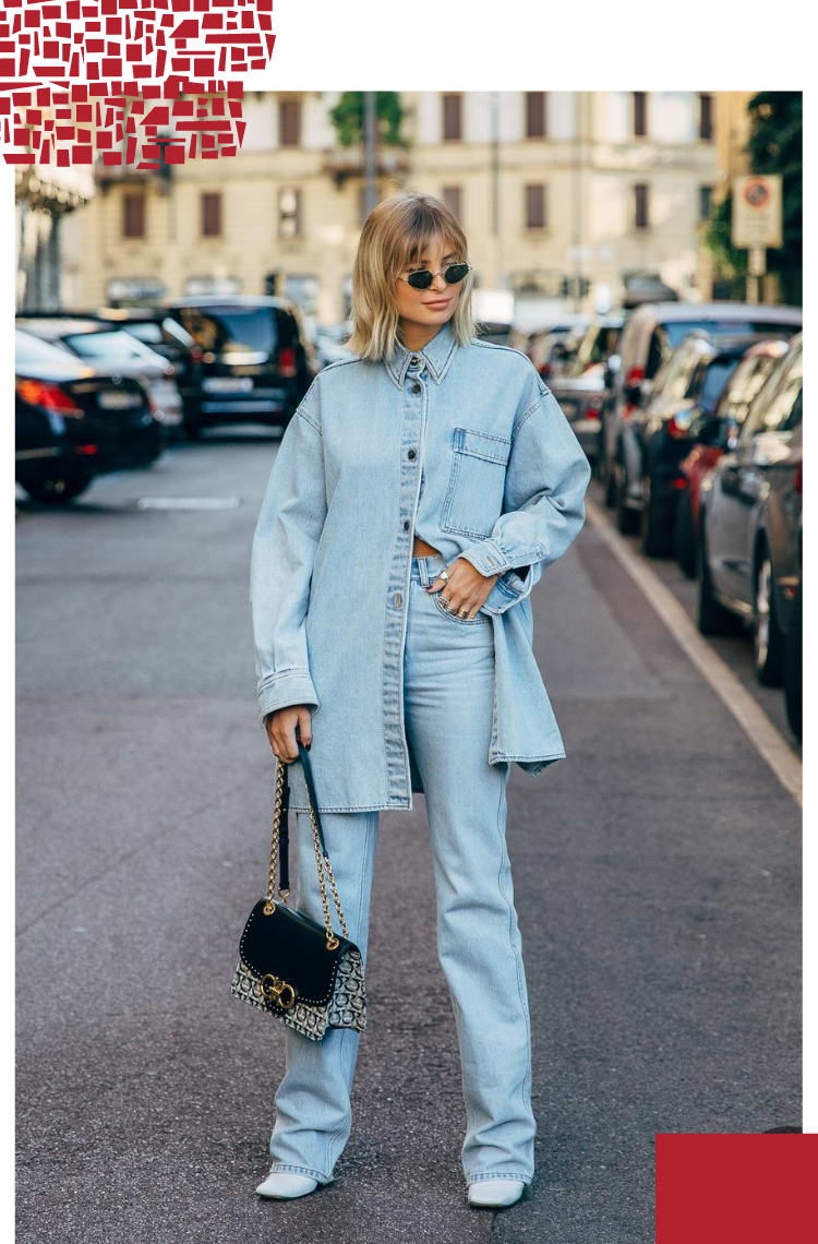 Foto vertical com moldura branca.Na foto, uma mulher loira com franja e comprimento na altura do ombro está parada em um estacionamento com muitos carros. O look é composto por uma jaqueta jeans de lavagem clara com botões de baixo abertos, calça jeans na mesma cor da parte de cima, bolsa preta, com alça de corrente dourada e monograma da Gucci e sapato branco.