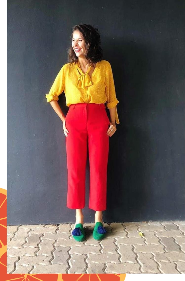 Foto vertical com moldura branca fina. Na foto, Marcie posa em frente a uma parede preta sorrindo e olhando para o lado sorrindo. O look é composto por camisa amarela com gola laço e estampa poá branca, calça vermelha de alfaiataria com barra curta e mule rasteiro verde com pompom azul.
