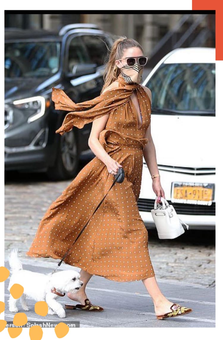 Foto vertical com borda branca e detalhe laranja. no topo direito. Na imagem, Olivia Palermo caminha na rua com seu cachorro em uma guia. O look é composto por vestido mídi acinturado com decote v e lenço ao redor do pescoço marrom com poá branco, lenço de seda bege amarrado no rosto, óculos escuros com armação clara e bolsa branca.