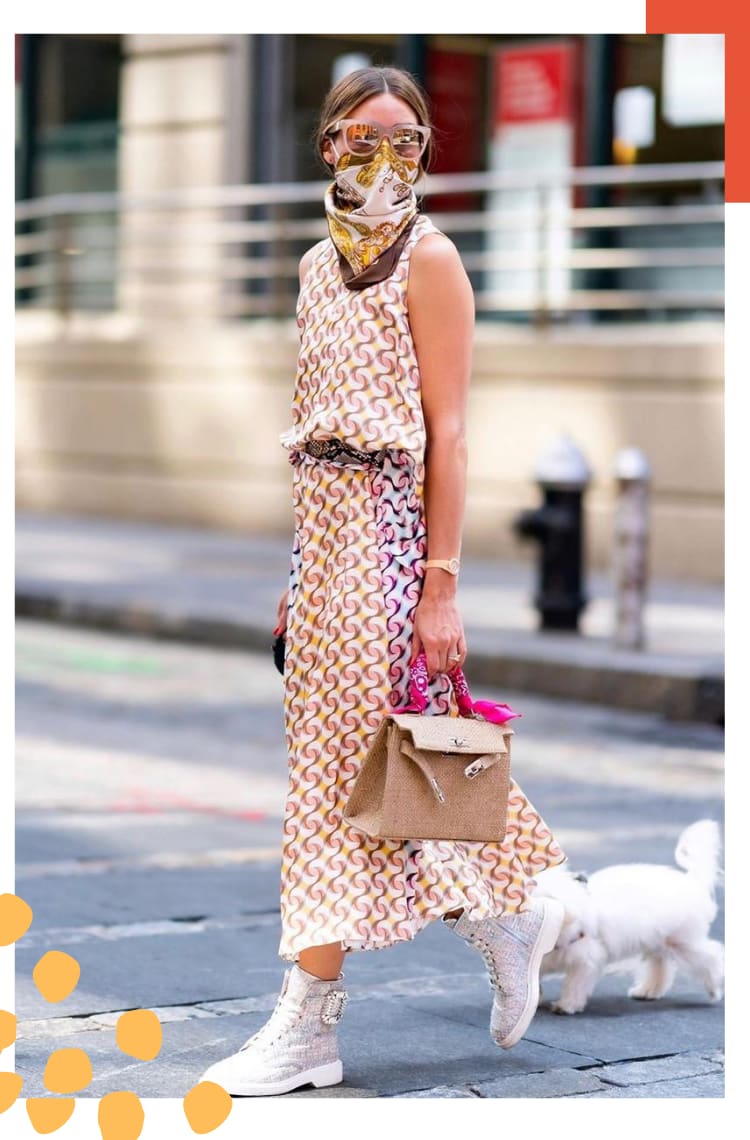 Foto vertical com borda branca e detalhe laranja. no topo direito. Na imagem, Olivia Palermo caminha na rua com seu cachorro em uma guia. O look é composto por um vestido mídi com estampa art noveau em tons de marrom, rosa e amarelo, lenço de seda com estampas douradas amarrado no rosto, óculos espelhado com armação bege e bolsa bege com um lenço rosa amarrado na alça.