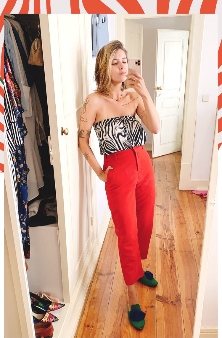 Foto com borda animal print laranja, mostrando como usar saia animal print em 8 looks. Na foto, Marcie tira foto em frente a um espelho, com celular na mão. O look é composto por saia de zebra preto e branco usada como blusa sem alças, calça de alfaiataria vermelha com barra curta e mule verde com pompom azul.