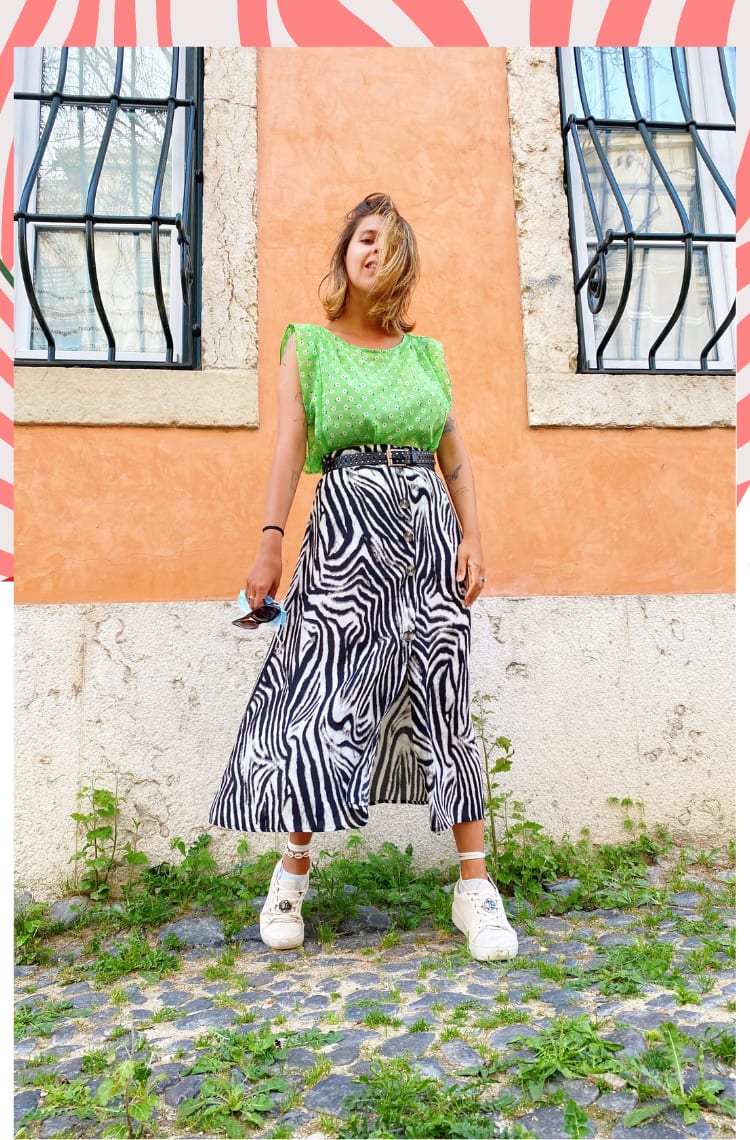 Foto com borda animal print laranja, mostrando como usar saia animal print em 8 looks. Na foto, Marcie, com cabelos loiros lisos na altura o ombro está em frente a um prédio laranja com janelas pretas ao fundo de ferro. O look é composto por regata com babados nos ombros verdes néon com estampa floral pequena branca, saia mídi fluída com botões frontais e estampa de zebra preta e branca, cinto preto com detalhes metálicos e tênis branco com cadarço amarrado na canela.