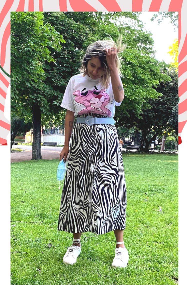 Foto com borda branca, mostrando uma forma de usar saia de zebra em looks. O look é composto por saia de zebra mídi com botões frontais, camiseta branca com estampa pantera cor de rosa, cinto azul claro e tênis branco.
