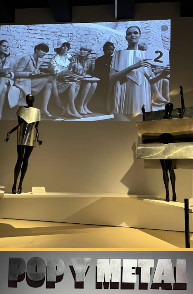 Um telão exibindo cenas do filme Quem é Polly Magoo? passam em uma parede branca. Embaixo, estão dois manequins pretos vestindo figurinos do filme, vestidos geométricos feitos de metal.
