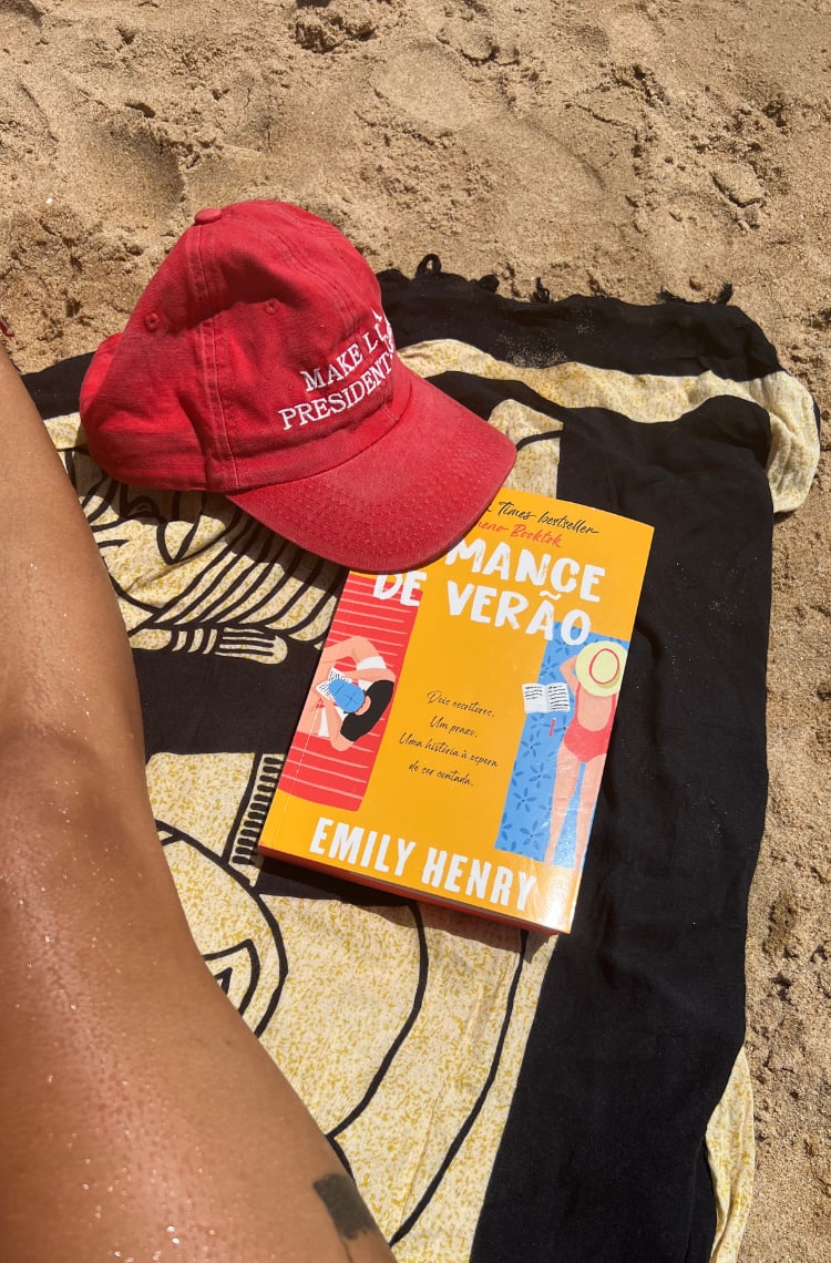 Foto de um dos 3 livros para ler no verão. A areia com uma canga preta com bege, um boné vermelho com um bordado branco escrito Make Lula President Again e um livro chamado Romance de Verão, com capa amarela e um homem e uma mulher lendo em suas cangas, em lados opostos da capa.