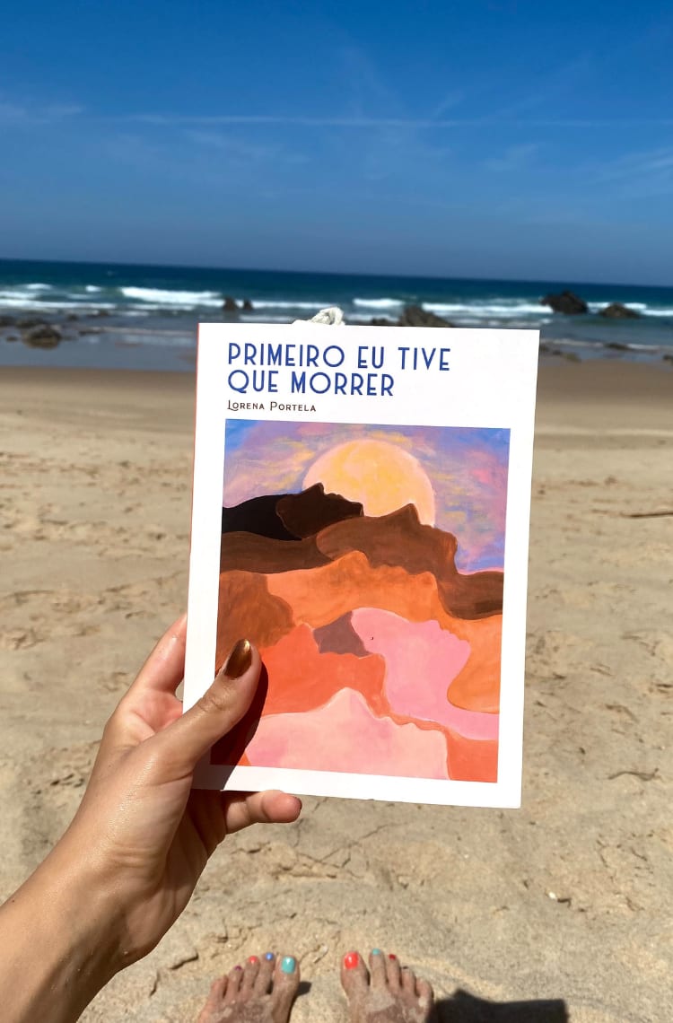 Foto de um dos 3 livros para ler no verão. Um livro de capa branca com o título Primeiro eu tive que morrer, de Lorena Portela está no centro da foto, segurado por uma mão. Ao fundo, está o mar e areia.