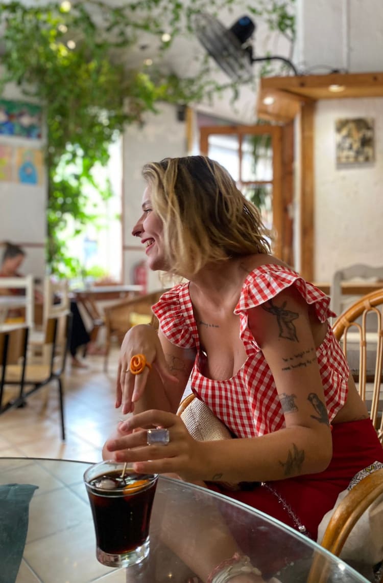 Foto para ilustrar uma das dicas 4 bebidas e pratos típicos de Valência. Marcie, uma mulher com cabelos curtos loiros e look vermelho está dentro de um bar, sorrindo para o lado, enquanto segura um copo de vermut.