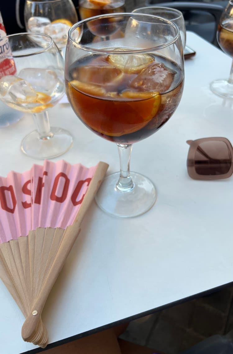 Foto para ilustrar uma das dicas 4 bebidas e pratos típicos de Valência. Foto para uma mesa branca está com taças de vidro cheia de vermut. Ao fundo estão um leque rosa e óculos de sol quadrados, também em tom de rosa.