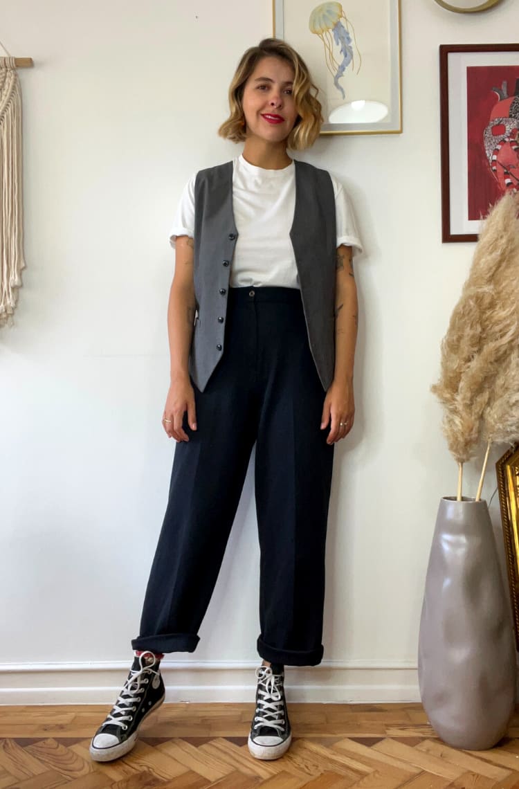 Foto mostrando a dica 1 de looks com colete. Marcie, criadora do blog, posa em frente a uma parede branca com quadros coloridos. O look é composto por camiseta branca usada por baixo de um colete cinza de alfaiataria usado aberto, calça preta de alfaiataria com a barra dobrada e tênis all star preto botinha.