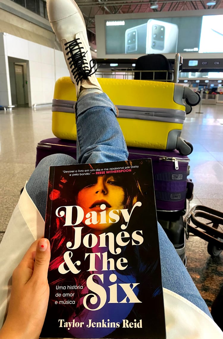Foto com o livro Daisy Jones & The Six, classificado como o terceiro melhor livro de Taylor Jenkins Reid por este blog. O livro está no centro da imagem e ao fundo estão algumas malas coloridas e um aeroporto desfocado ao fundo. A capa conta com o título e a foto colorida em tons de amarelo, roxo e vermelho de uma mulher com cabelos longos e franja.