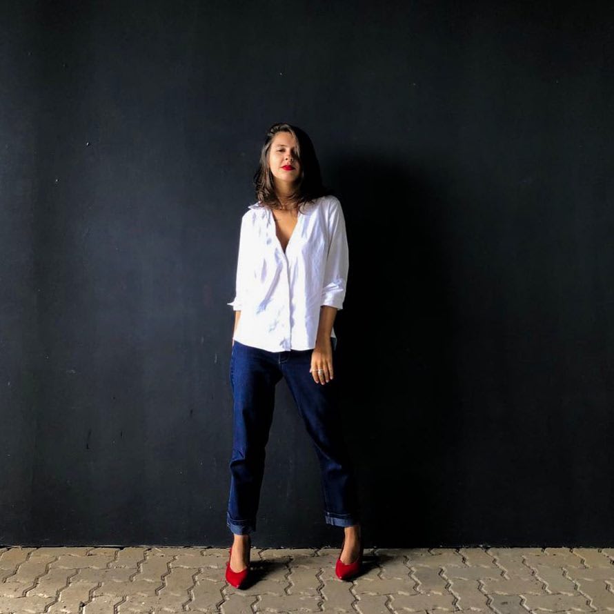 Marcie posa em frente a uma parede preta olhando para a câmera. O look é composto por camisa branca solta, calça de alfaiataria boyfriend com barra dobrada e scarpin vermelho.