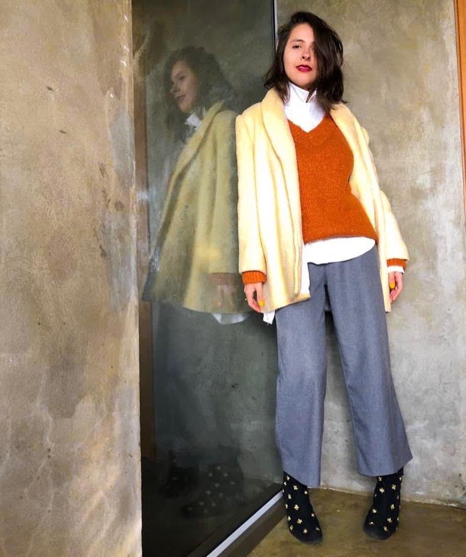 Marcie posa em frente a uma parede cinza com vidro. O look é composto por camisa branca usada por baixo de um blusão mostarda com gola v, casaco amarelo de lã, pantacourt cinza de lã e bota preta com bordado dourado.