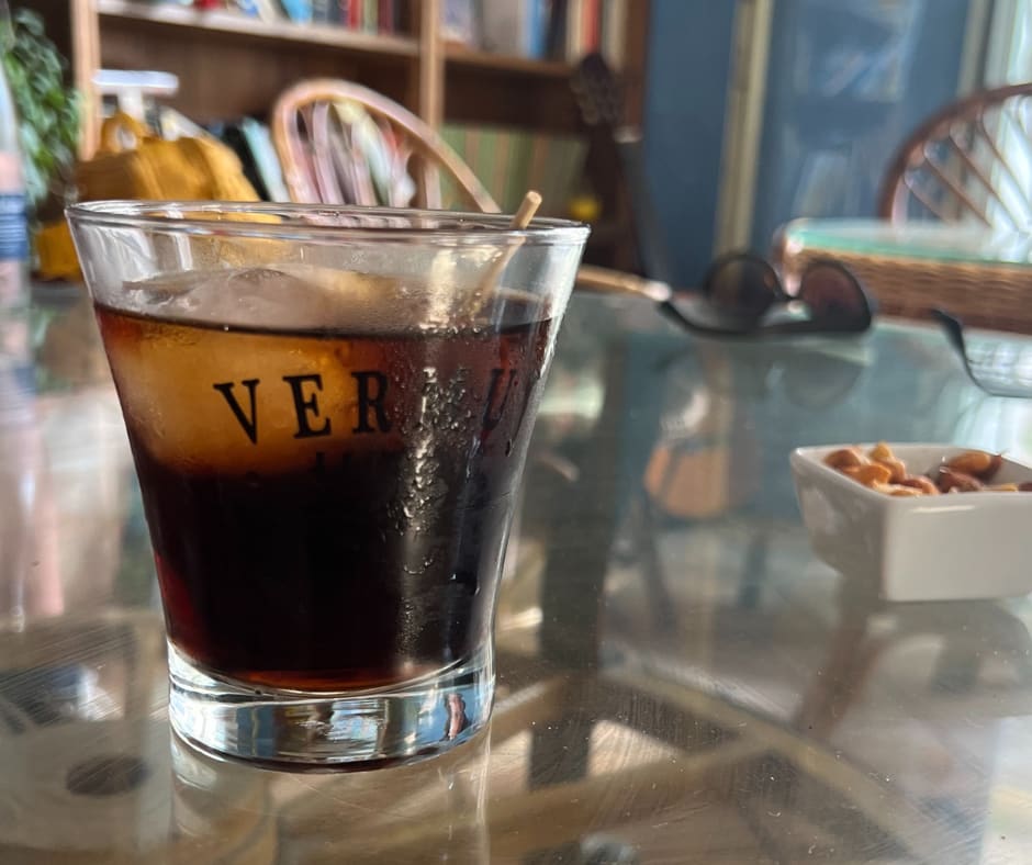 Foto de abertura do post com dicas. de 4 bebidas e pratos típicos de Valência. Uma mesa transparente está com um copo de aperitivo de vermut cheio e um pote quadrado de amendoins.