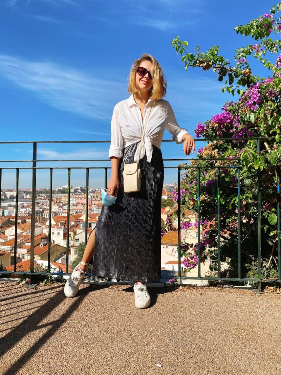 Marcie posa em frente a um miradouro, com céu azul e mini casas ao fundo. O look é composto por camisa branca amarrada na cintura, saia longa prateada, bolsa branca a tiracolo e tênis branco com cadarços amarrados na canela.