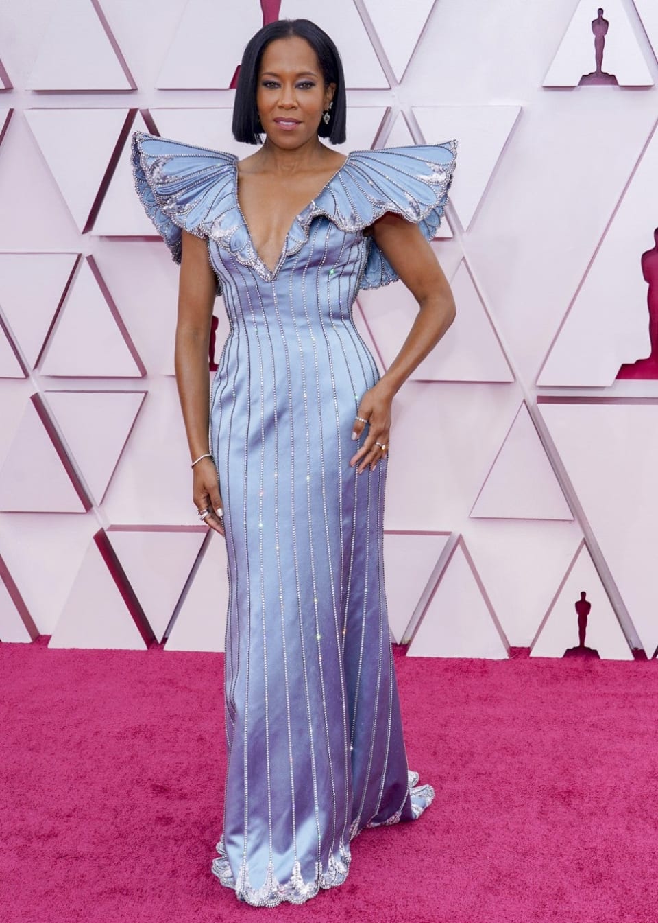 Foto do look de Regina King no red carpet do Oscar 2021. Ela veste um vestido azul claro acetinado longo com decote v profundo e mangas amplas altas, que parecem asas de borboletas. O vestido tem linhas verticais feitas com cristaias e lantejoulas. O cabelo está liso na altura dos ombros, com brincos discretos de cristais.