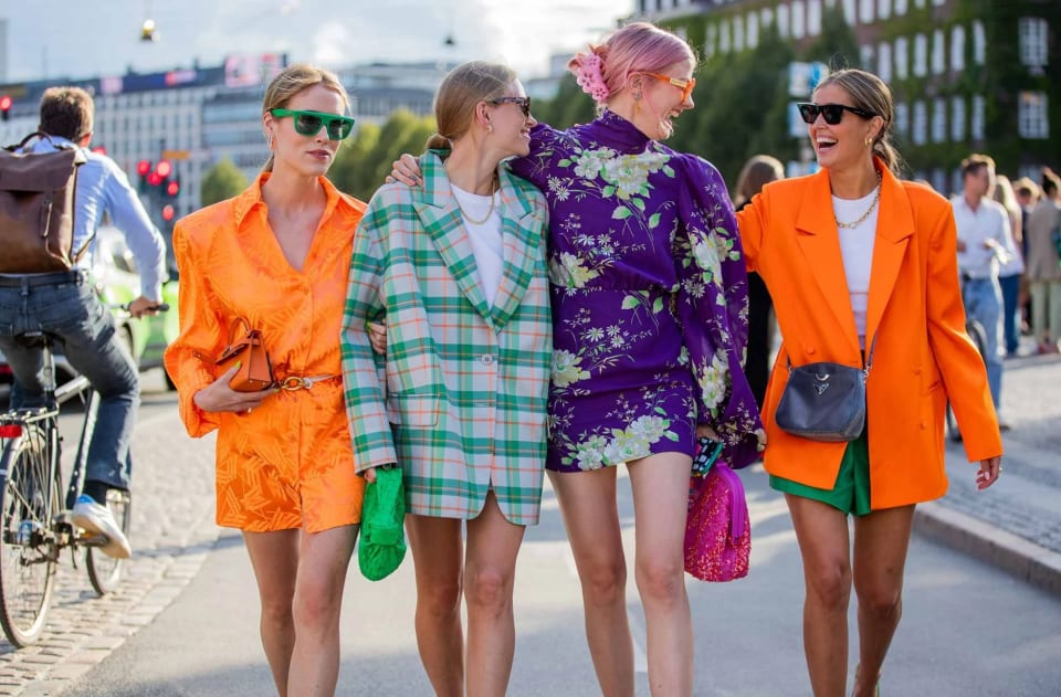 Foto com quatro amigas abraçadas em uma rua movimentada com pessoas desfocadas ao fundo. O look da primeira é composto por camisa laranja usada como vestido, cinto dourado, bolsa laranja míni e óculos escuros com armação verde. A segunda veste um blazer oversized xadrez verde com laranja, usado por cima de uma camiseta branca, bolsa verde e colar dourado. A terceira veste um míni vestido de mangas longas roxa com estampa floral verde e bolsa rosa na mão. A quarta veste uma camiseta branca com short verde, blazer laranja e bolsa cinza.