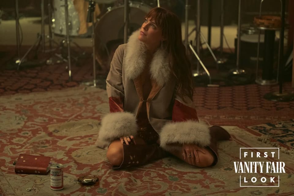 Foto da atriz que interpreta Daisy Jones sentada no tapete de um estúdio. O look é composto por casaco de pelos e bota de cano alto.