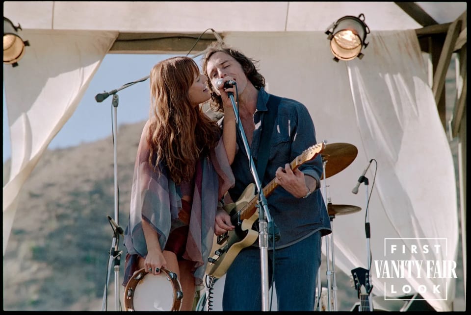Foto do elenco de Daisy Jones & The Six em cima de um carro. Os protagonistas da série, Daisy Jones e Billy estão dividindo um microfone em um palco de festival, com tecidos brancos esvoaçantes no palco. Ela usa um quimono multicolorido e ele veste um look de camisa e calça jeans azul.