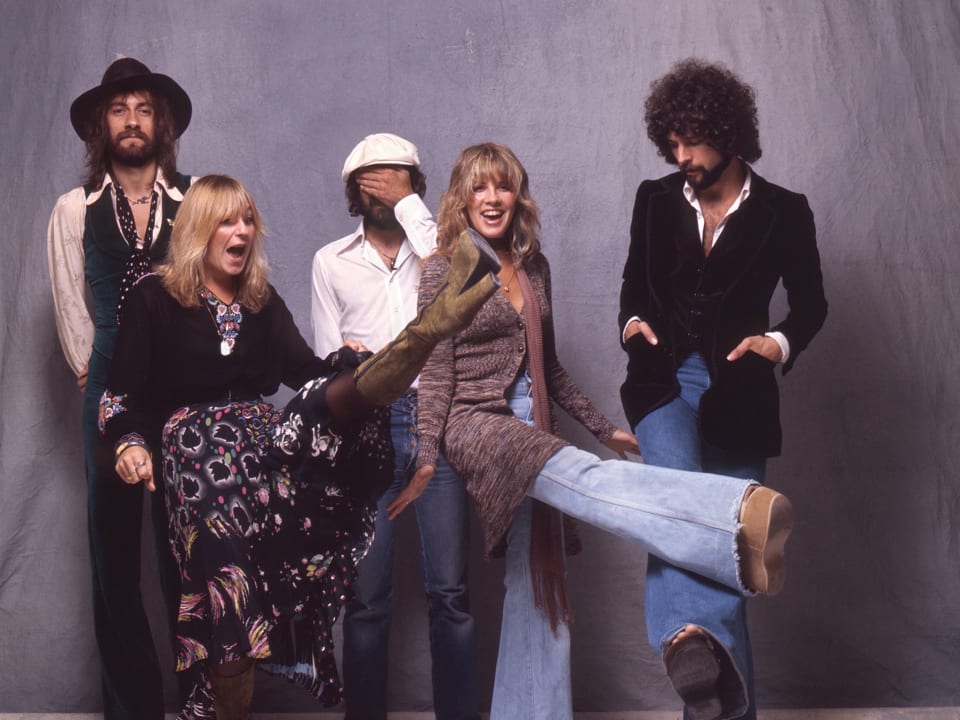 Foto do grupo Fleetwood Mac, banda que inspira o livro Daisy Jones & The Six. Os 5 membros da banda estão lado a lado em um fundo cinza, usando looks de estilo boho.