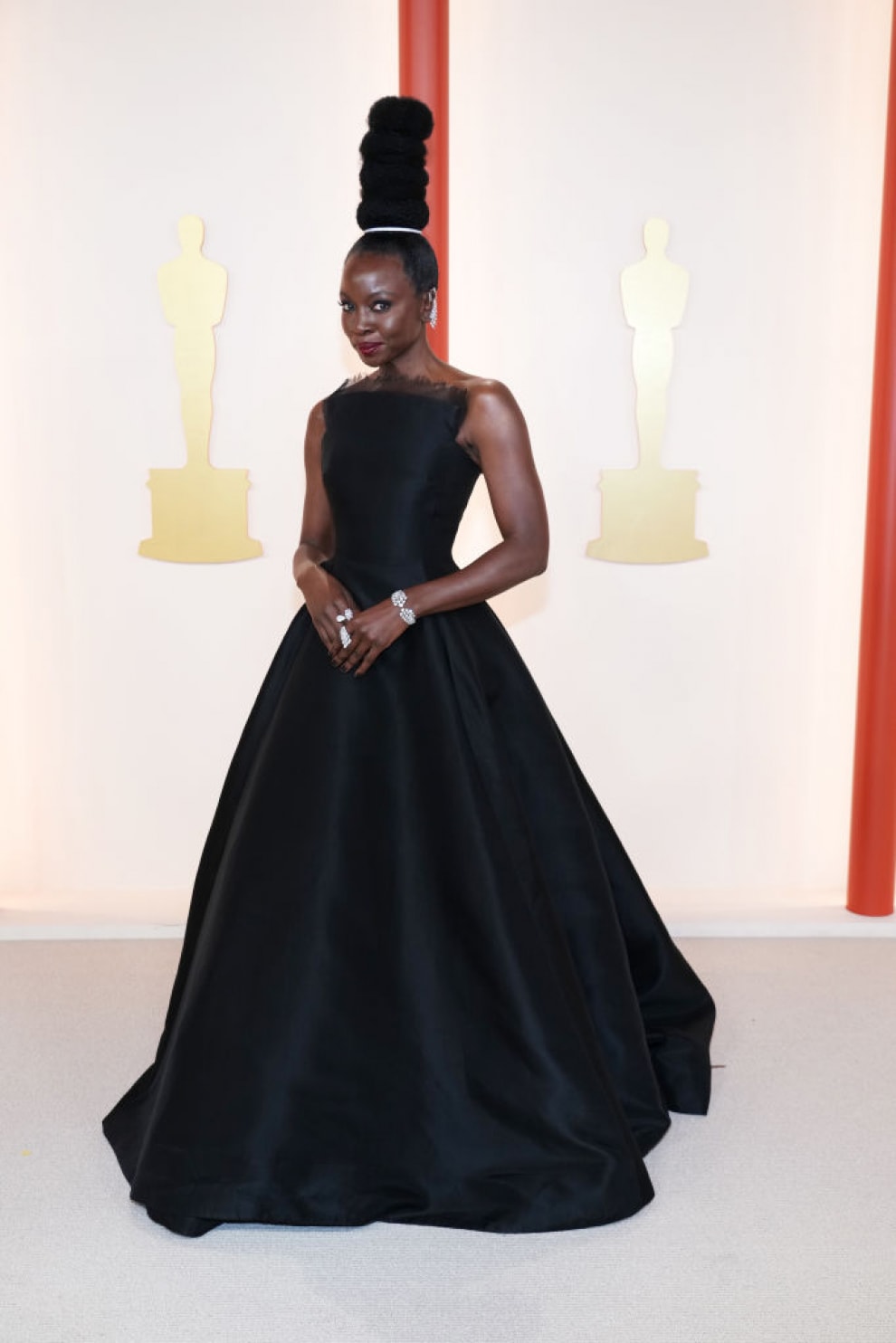 Foto ilustrando os melhores looks do oscar 2023. Na foto, a atriz Danai Gurira está no tapete vermelho. O look é composto por um vestido longo sem mangas preto com saia volumosa de baile, enquanto o penteado é uma escultura com quatro coques verticais.