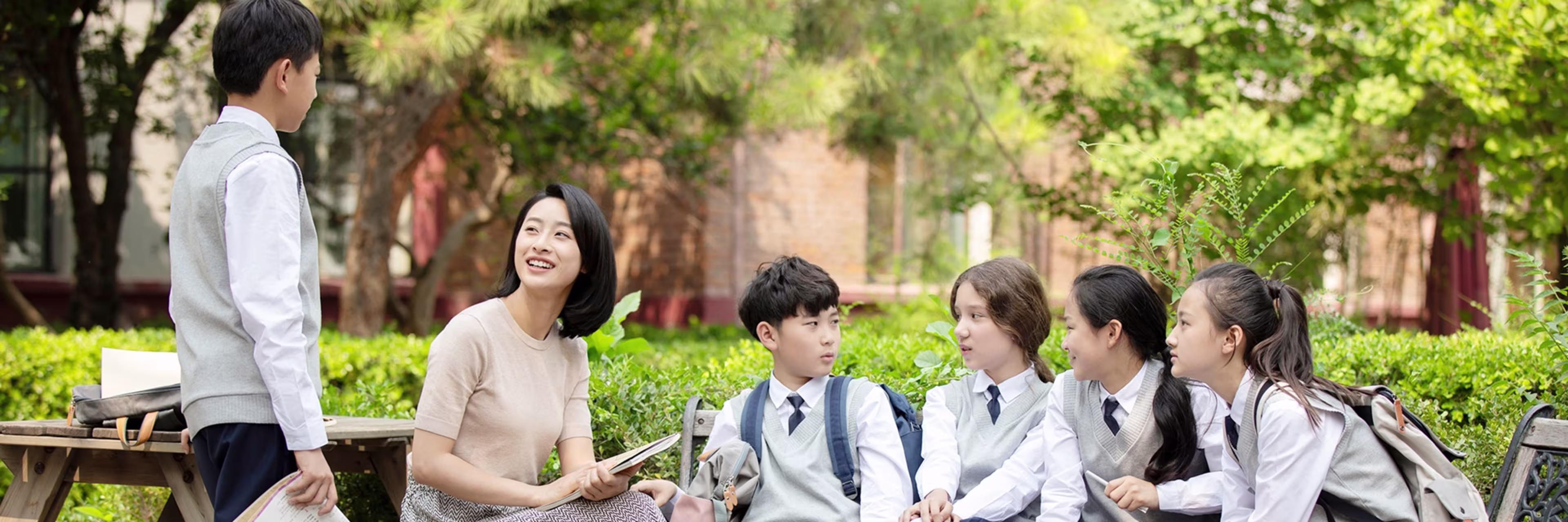 Teacher talking with students in school uniforms on a bench in a green courtyard.