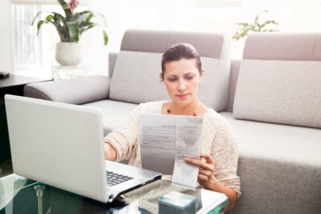 woman sitting in front of couch, checking document and using her laptop