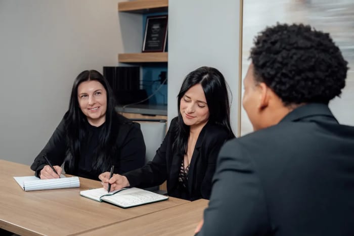 Two women taking notes at a conference table, facing a third person