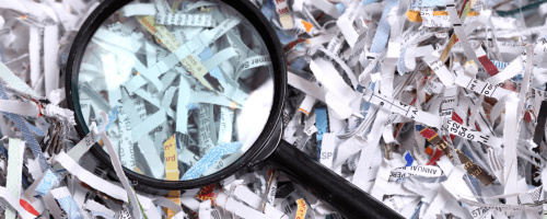 A magnifying glass lays across a pile of shredded papers. The image conveys the unsettling idea that even with precautions taken, anyone can become an identity theft victim. In the context of the page, the image implies that working with an identity theft lawyer can be critical to identity theft recovery.