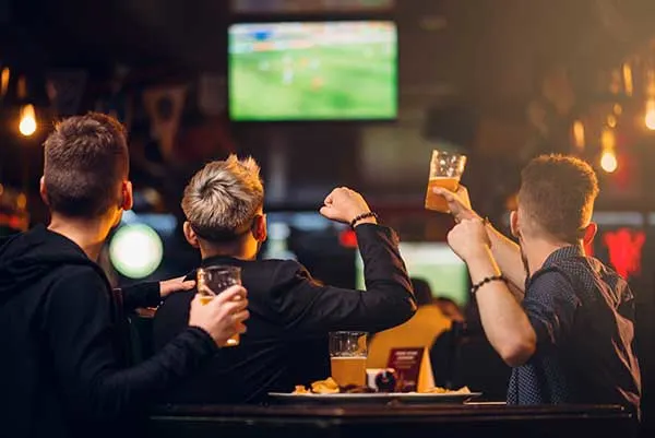men at sports bar, raising their beer glasses and cheering while watching a game on a television screen