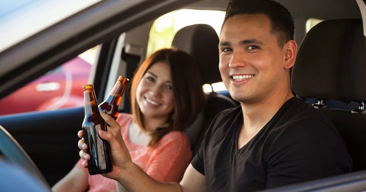 A smiling man and woman sitting in a car, each holding a beer bottle, with the man in the driver's seat.
