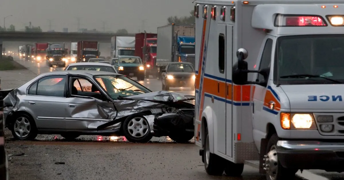 A silver car, heavily damaged, sits on a rain-slicked highway next to an ambulance. Other vehicles are visible in the background under a cloudy sky.