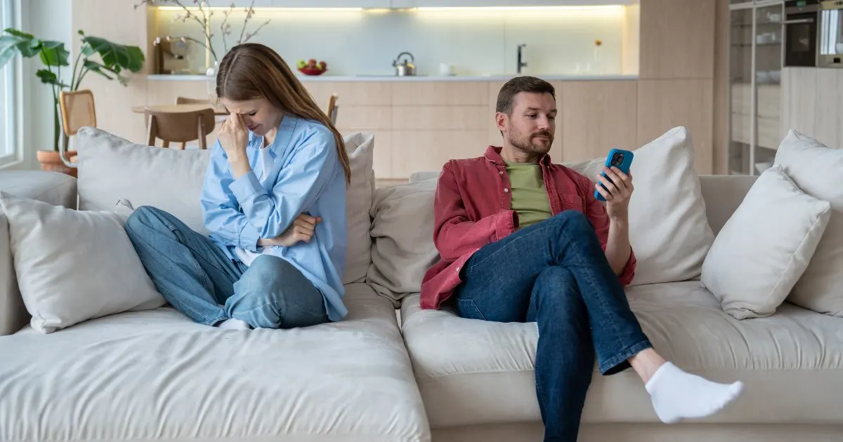 A couple sits on a couch, separated by a large gap. The woman on the left is crying, while the man on the right ignores her, looking at his phone.