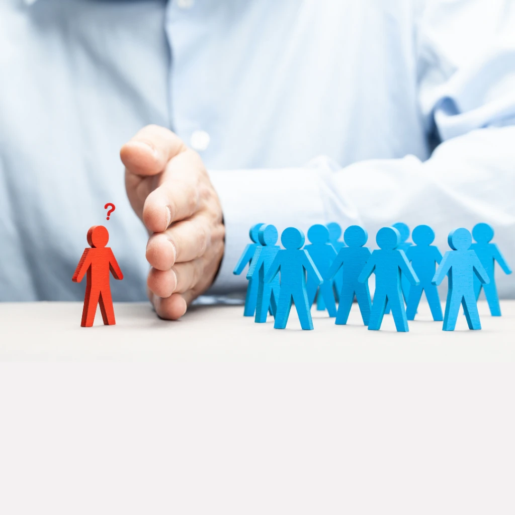 Only the torso and arm of a man wearing a button down shirt are visible. His hand is positioned on a desktop vertically, with his pinky finger on the bottom, pressed into the desktop and his thumb at the top. His hand is intentionally forming a barrier between a group of people (represented as little blue 3D cutouts standing around in a group) and a single person (shaped as a little red 3D cutout standing by itself). The red person has a small red question mark above their head as if they can't understand why the man is excluding them from the group. The image symbolizes the uncertainty that some people feel about whether discrimination is the root cause of their workplace situation and whether they need to speak with a discrimination lawyer.