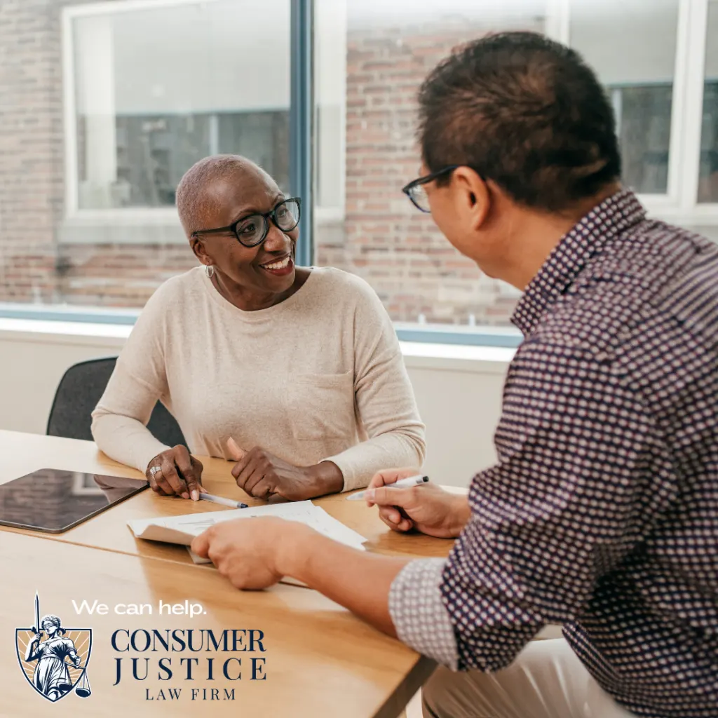 A client works with an attorney in a conference room, with papers on the table between them. Large windows behind the client reveal a brick building in the background. The client and attorney are facing each other, smiling. Only the back and side profile of the attorney is visible. The attorney is helping his client with her HireRight background check errors.