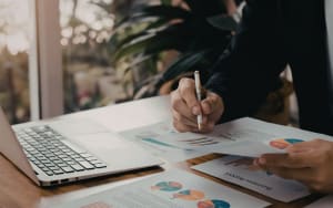 A person holding a pen and reviewing business reports with charts and graphs next to a laptop on a wooden desk.