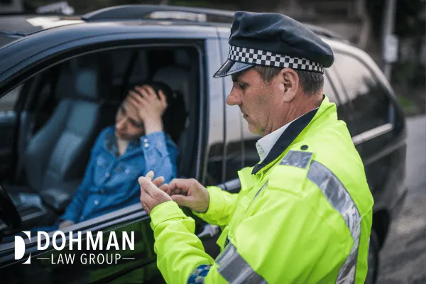 Woman in a car with her hand on her head while a police office is outside her window looking at her driver's license. She is wondering "What Happens if you get a Ticket on an International License?"